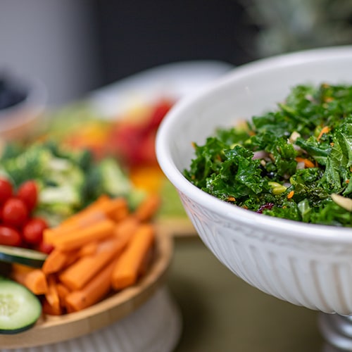 Bowl of healthy greens with a plate of carrot sticks, cherry tomatoes, cucumbers, and broccoli